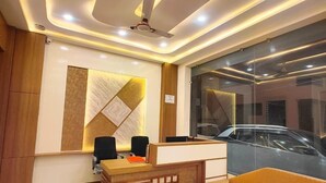A modern reception area with a reception desk, two black office chairs, a sofa, and a large window with a view of a car outside. - Hotel Vistara (Ujjain)