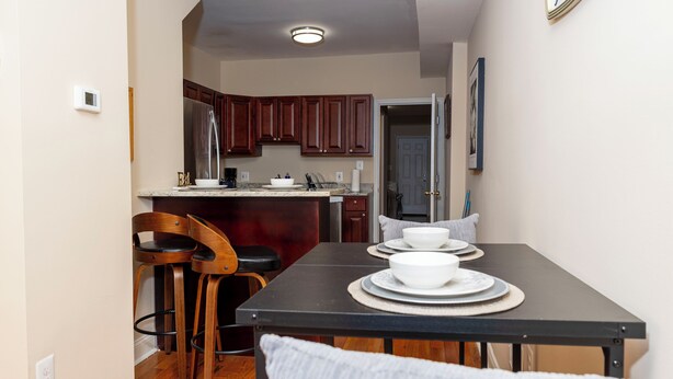 A compact kitchen with wooden cabinets, a dining table with white plates, and a bar stool. - 3BR•4 Bath•2-Min Walk to Johns Hopkins Hospital. (Baltimore)