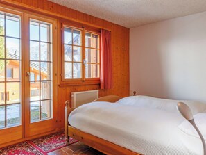 Ein Schlafzimmer mit einem hölzernen Bett, einem Heizkörper, einem Fenster mit Vorhang und Blick auf ein Gebäude.
