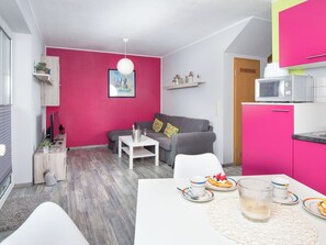 Un espacio habitable compacto con kitchenette, un sofá gris, una mesa blanca con productos para el desayuno y una pared rosa vibrante.
