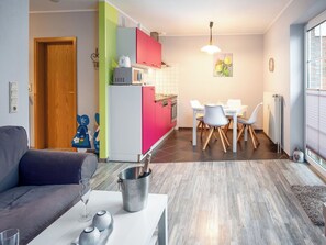 Una sala de estar moderna con un sofá gris, un comedor con mesa y sillas blancas, y una cocina con armarios de colores.