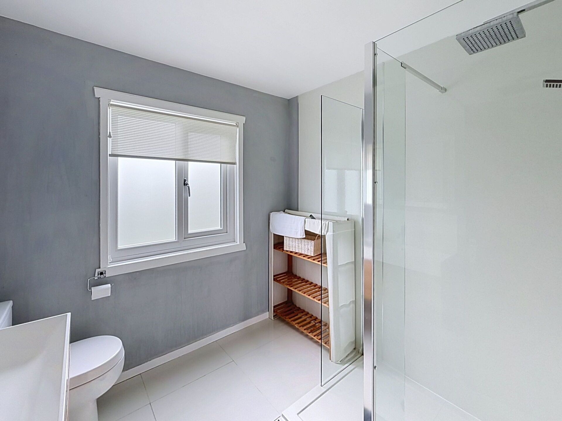 A bathroom with a toilet, a window with blinds, a glass shower enclosure, and a shelf with toiletries.