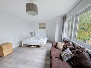Ein Schlafzimmer mit einem Bett, einer hölzernen Kommode, einem braunen Sofa, einem Fenster mit Blick und einer Pendelleuchte.