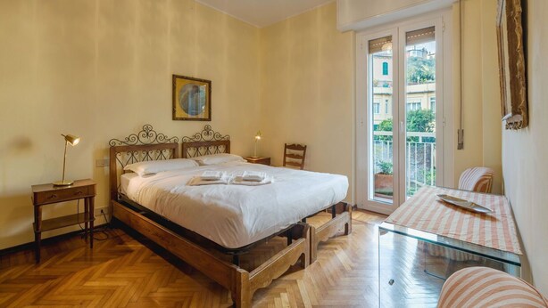 Une chambre à coucher avec un lit en bois, des tables de chevet, une table en verre et une vue sur les immeubles par la fenêtre.