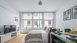 Une chambre moderne avec une grande fenêtre, un lit recouvert d’une couette grise, une télévision et une table où est disposé un petit-déjeuner composé de croissants et de jus d’orange.