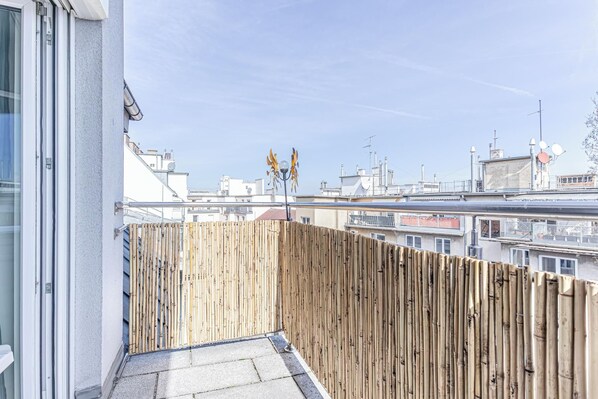Un balcon avec une balustrade en bois, une porte vitrée et une vue sur des immeubles et des antennes en arrière-plan.