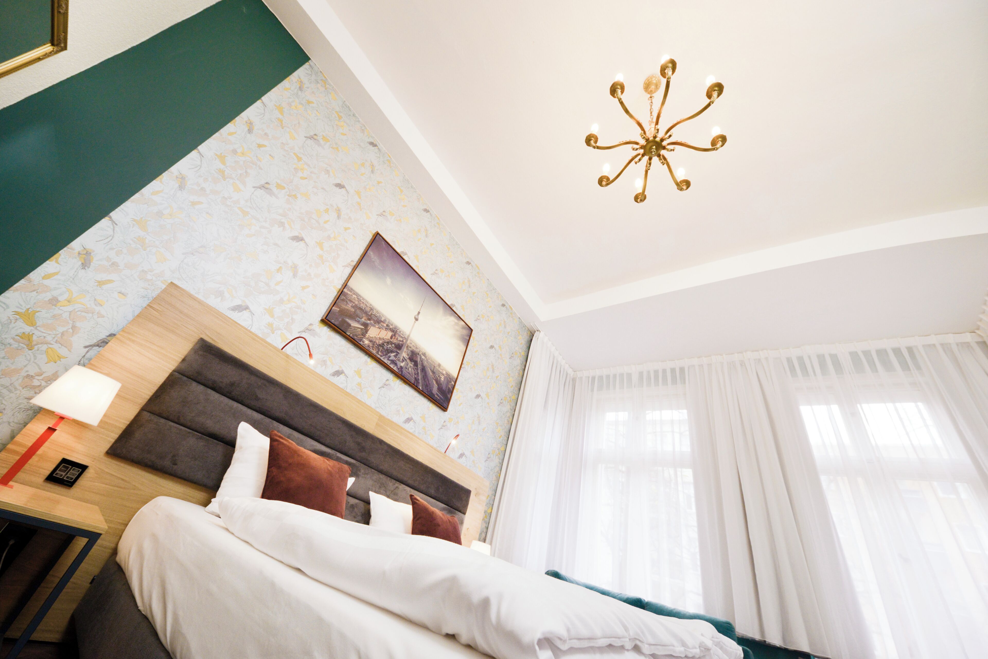 Hotellihuone, jossa on sänky, yöpöytä, lamppu, taulu ja kattokruunu.