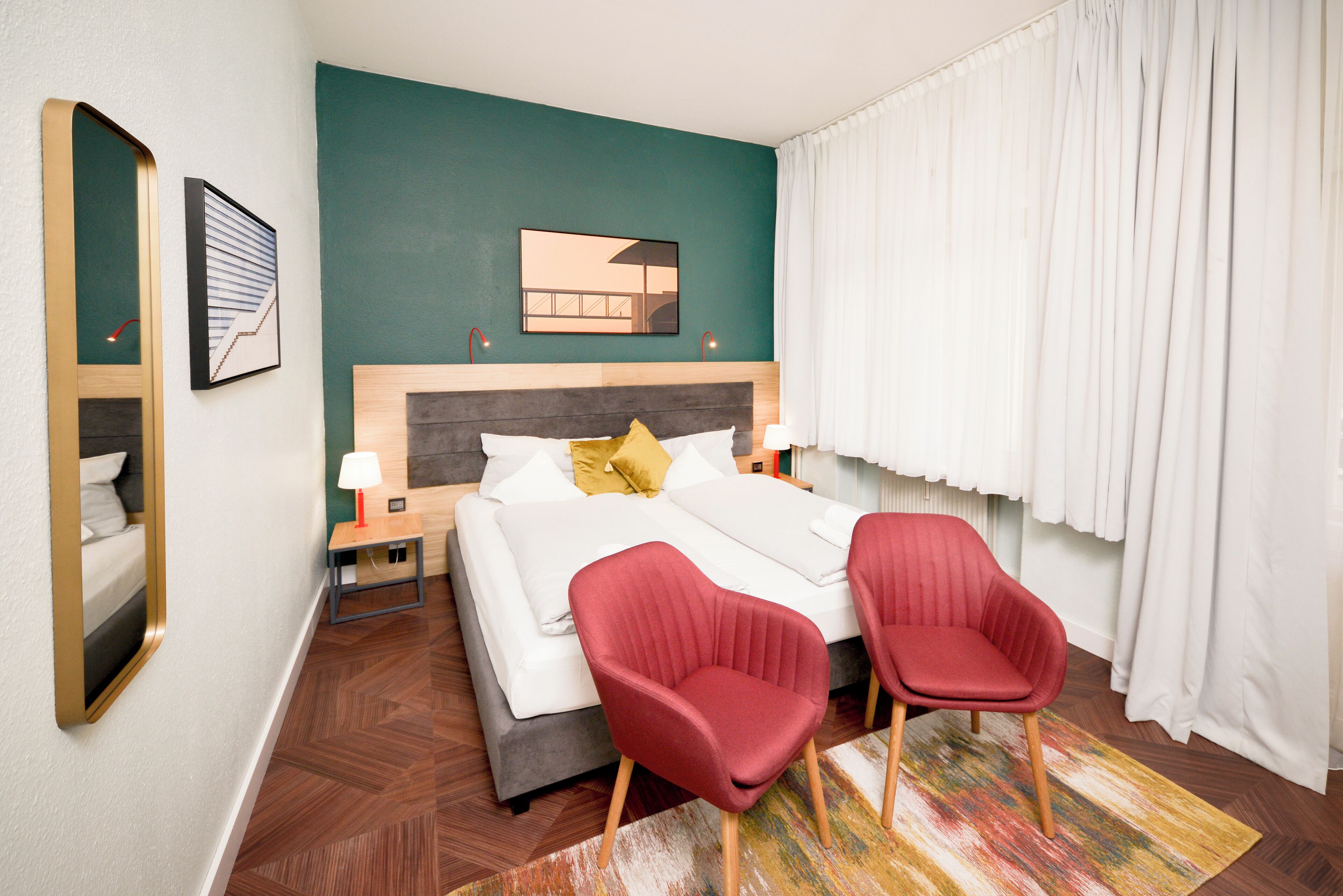 A hotel room with a bed, two red chairs, a nightstand, a lamp, and a mirror on the wall.