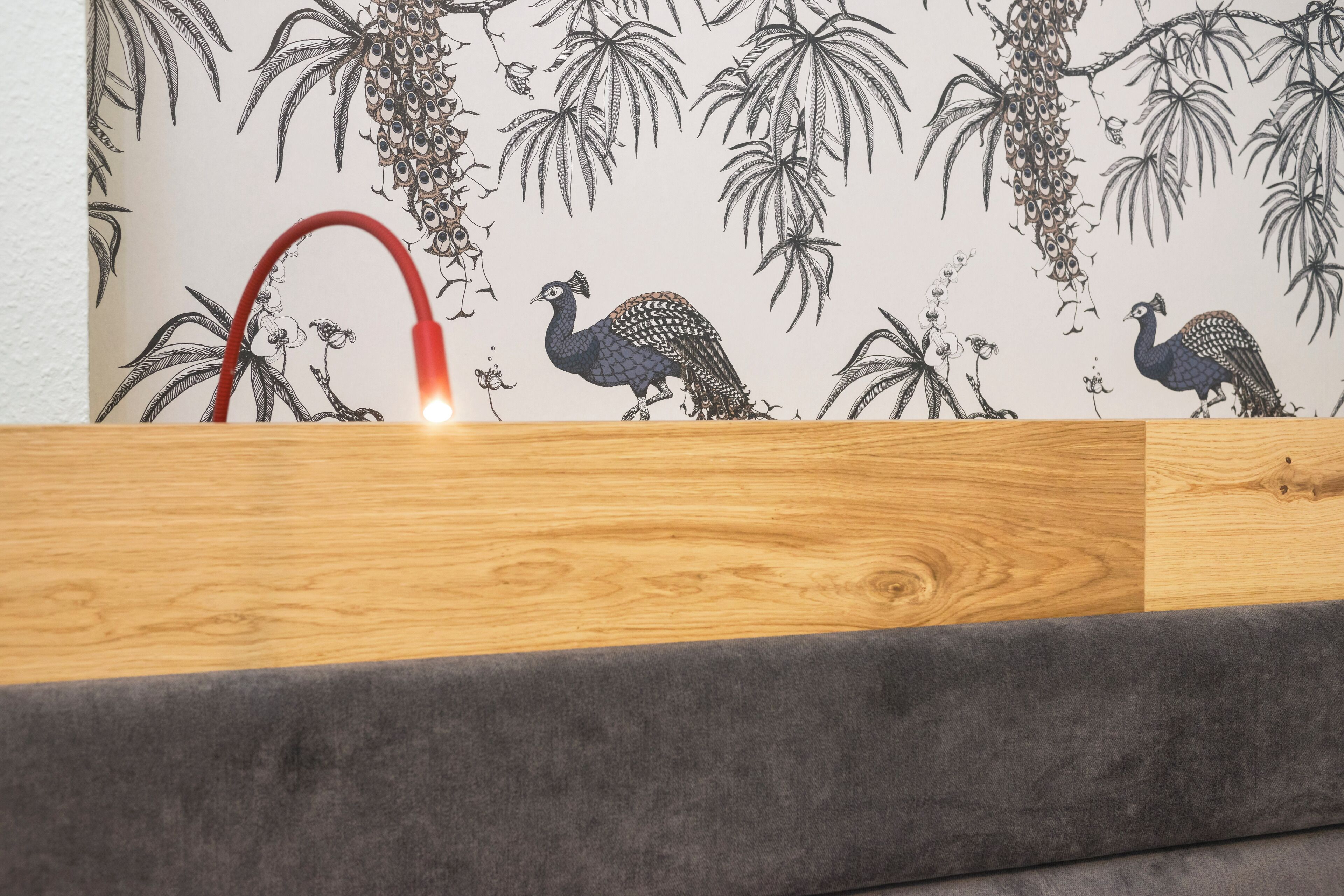 Ein Holznachttisch mit einer grau gepolsterten Bank vor einer Wand mit Tapete mit Pfauhen- und Pflanzenmuster.