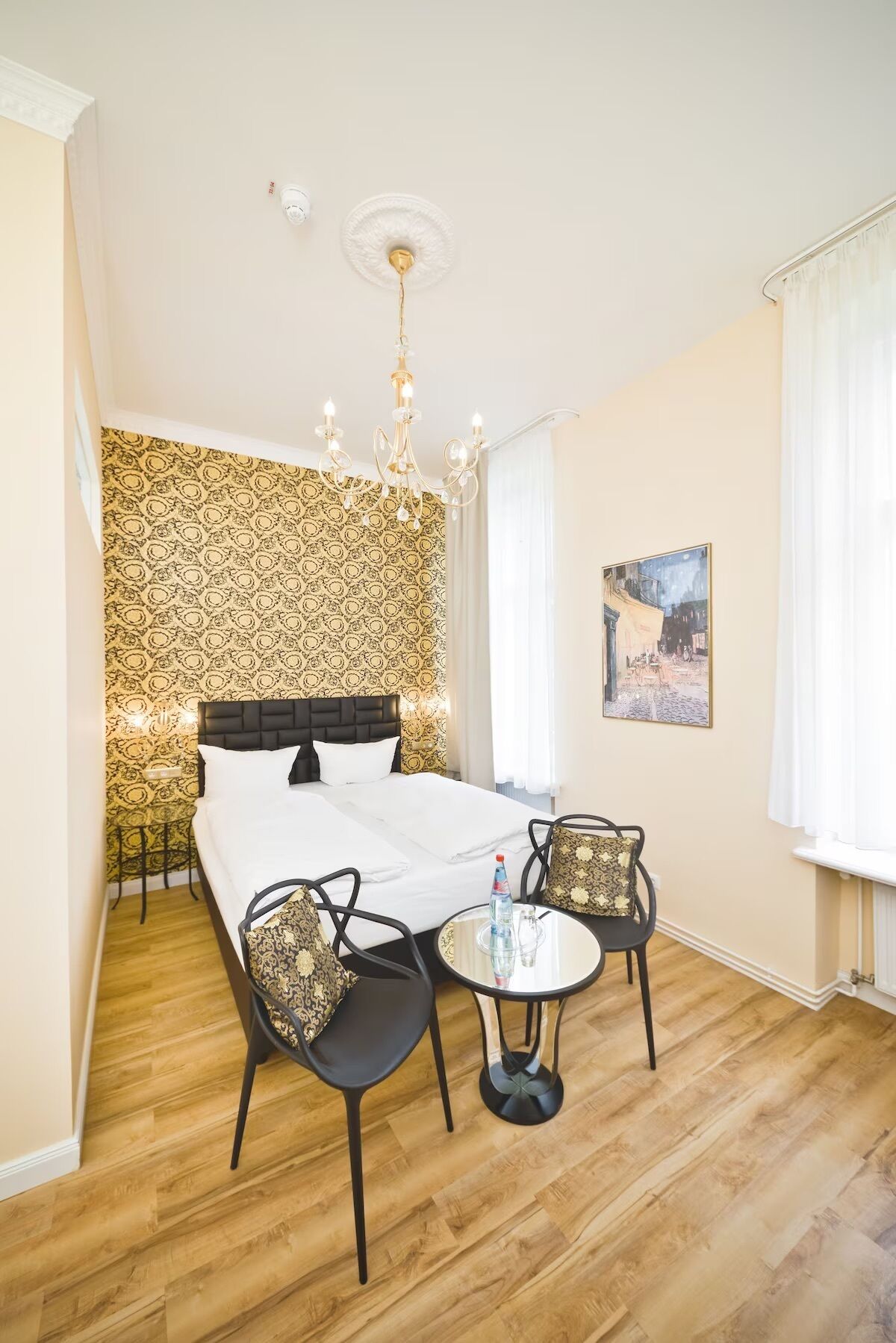 Une chambre d’hôtel avec un grand lit, un lustre, une petite table avec des chaises et un papier peint à motifs.