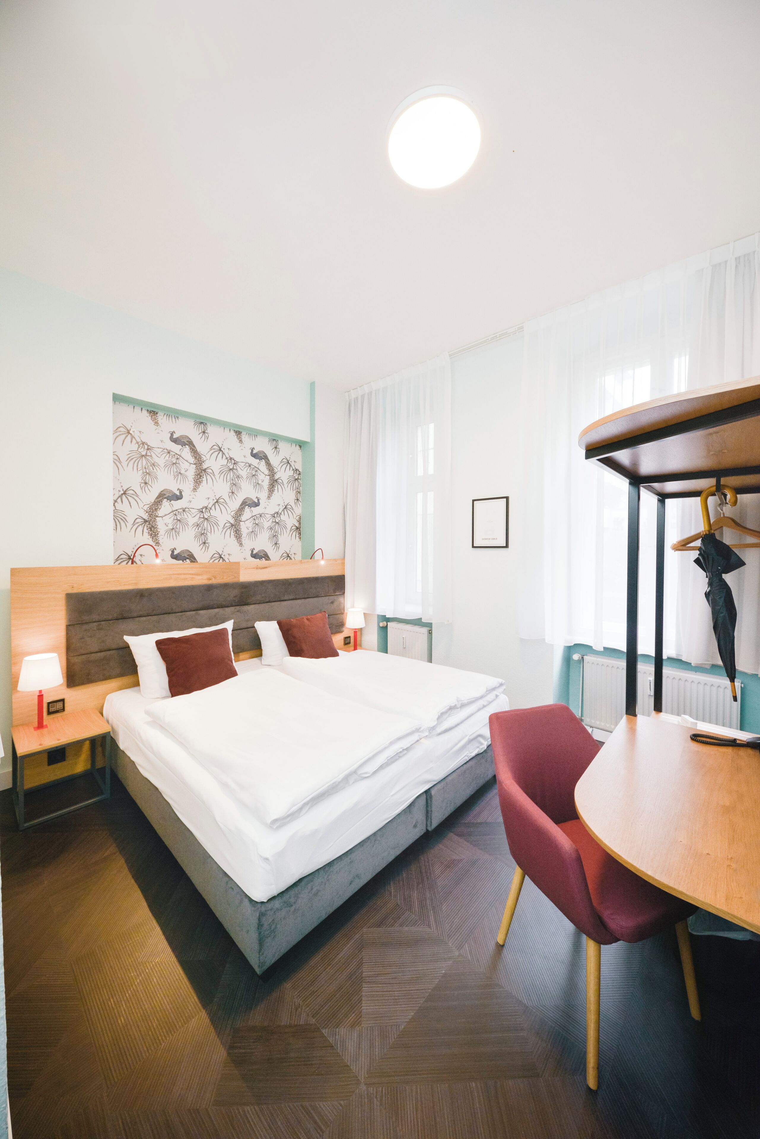 Hotellihuone, jossa on suuri sänky, puinen pääty, punainen tuoli, puinen pöytä ja seinälle ripustettu taideteos.