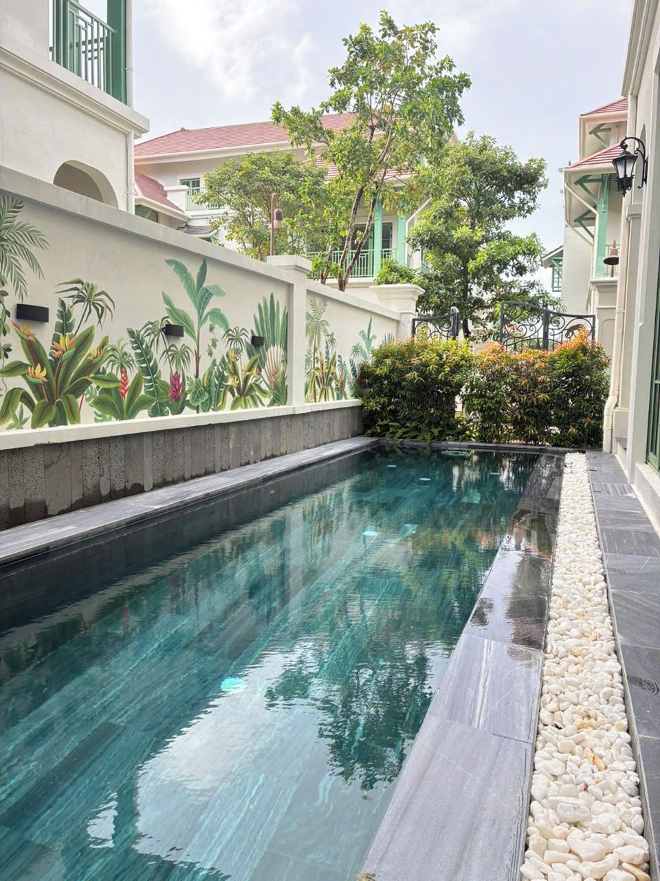 Piscina com superfície de água límpida, rodeada por um muro baixo com um mural tropical, e um caminho de pedra que leva à entrada de um edifício.