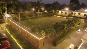 A well-lit outdoor area with a paved surface, surrounded by greenery and palm trees, featuring a stone wall with a sign and a tent structure. - Veer Garh Farms (Udaipur)