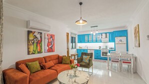 A living room with a sofa, a coffee table, and a dining area with a table and chairs. There is a kitchen in the background with blue cabinets and a white refrigerator. - Swim, Sauna, Sea: An Exclusive Forest Sanctuary with Mediterranean Panoramas (antalya)