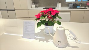 Una cocina moderna con armarios blancos, una encimera blanca, una tetera eléctrica blanca y una planta en maceta con flores rojas.