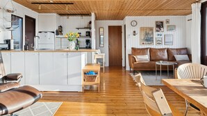Una cocina moderna con armarios blancos y una isla de madera, un área de estar con un sofá de cuero marrón y una mesa de comedor, y un techo de madera.