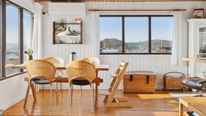 Una habitación con suelo de madera, una mesa de comedor con sillas, una cesta de mimbre y vistas al campo a través de grandes ventanales.