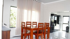 A dining area with a wooden table and chairs, a window with sheer curtains, and a kitchen area visible through an open door. - Tou Safari Lodge (Livingstone)