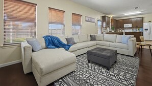 A living room with a sectional sofa, ottoman, patterned rug, and large windows with blinds.