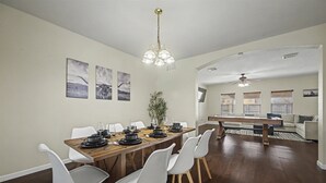 A dining area with a wooden table, white chairs, and a chandelier. There are framed pictures on the wall and a plant in the corner.