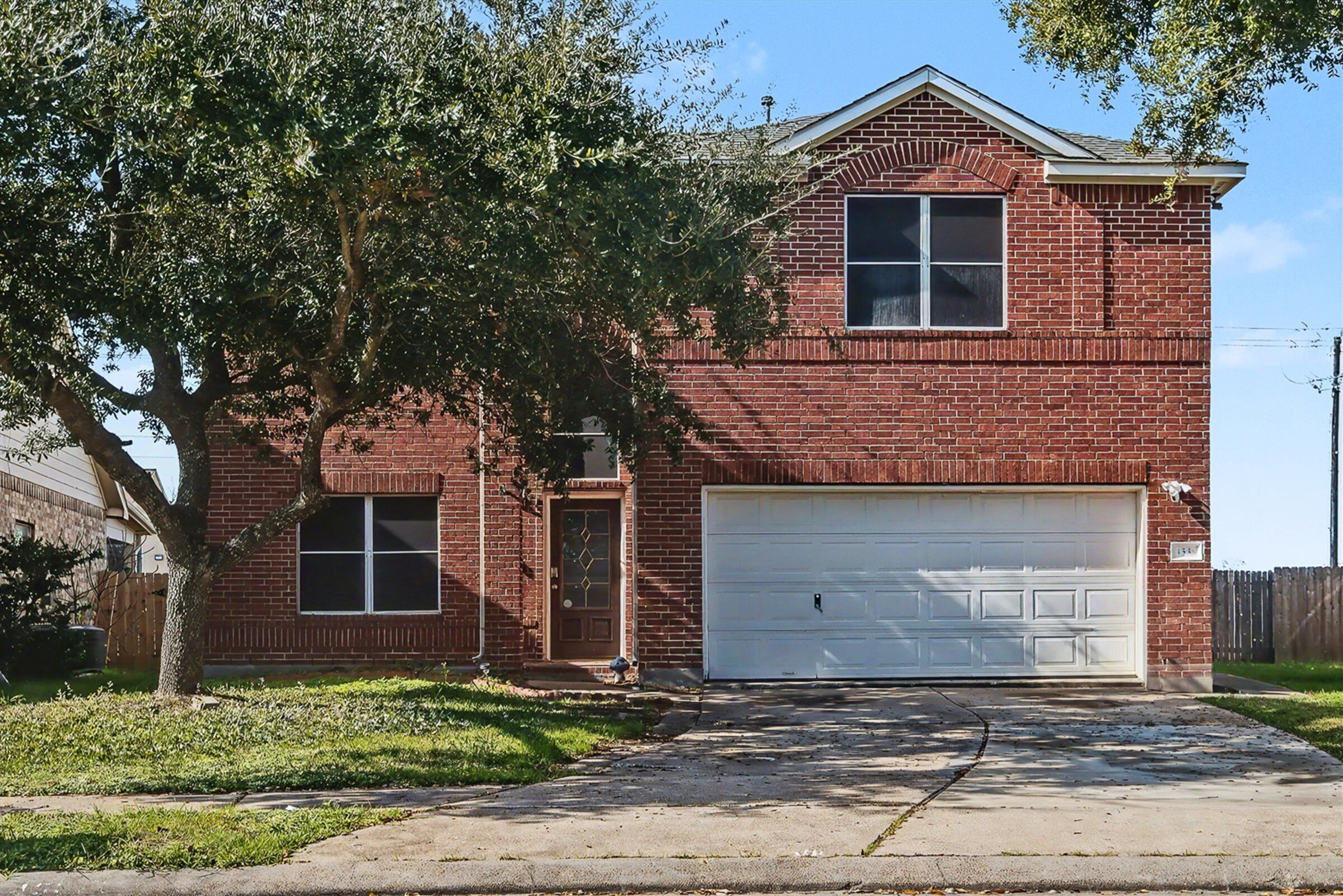 A two-story brick house with a white garage door, a front door, and a large tree in front.