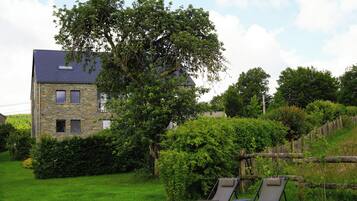 A stone building with a sloped roof, surrounded by lush greenery and a wooden fence.
