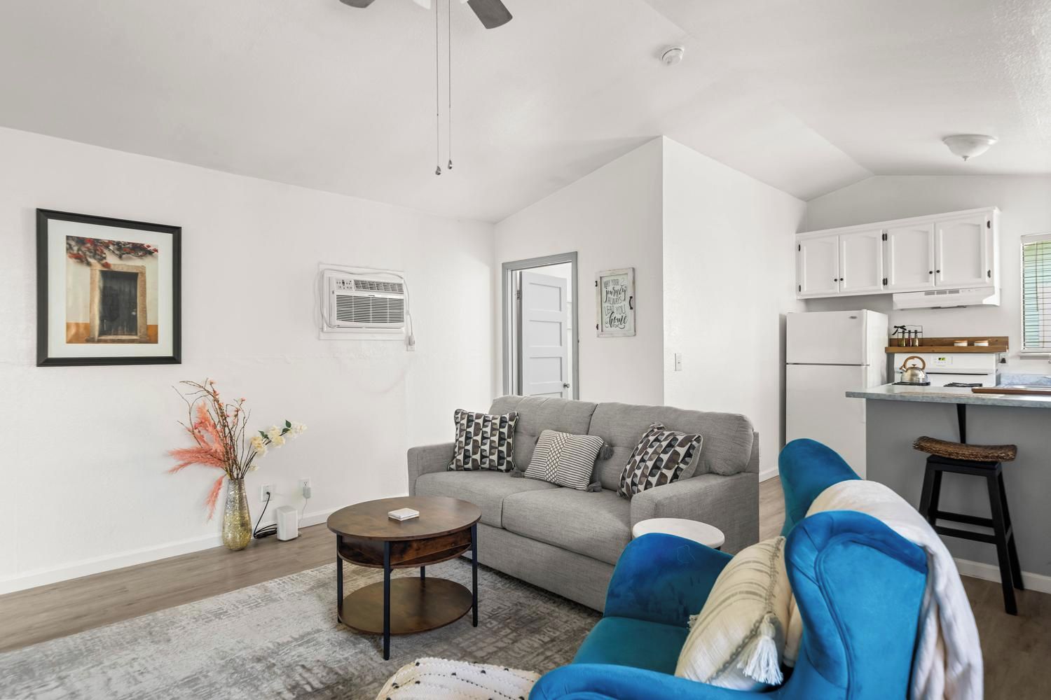 A modern living room with a grey sofa, a coffee table, and a blue armchair. There is a kitchen area with white cabinets and a countertop.
