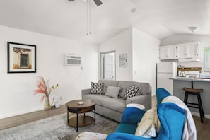Una sala moderna con un sofá gris, una mesa de centro y un sillón azul. Se observa una zona de cocina con armarios blancos y una encimera.