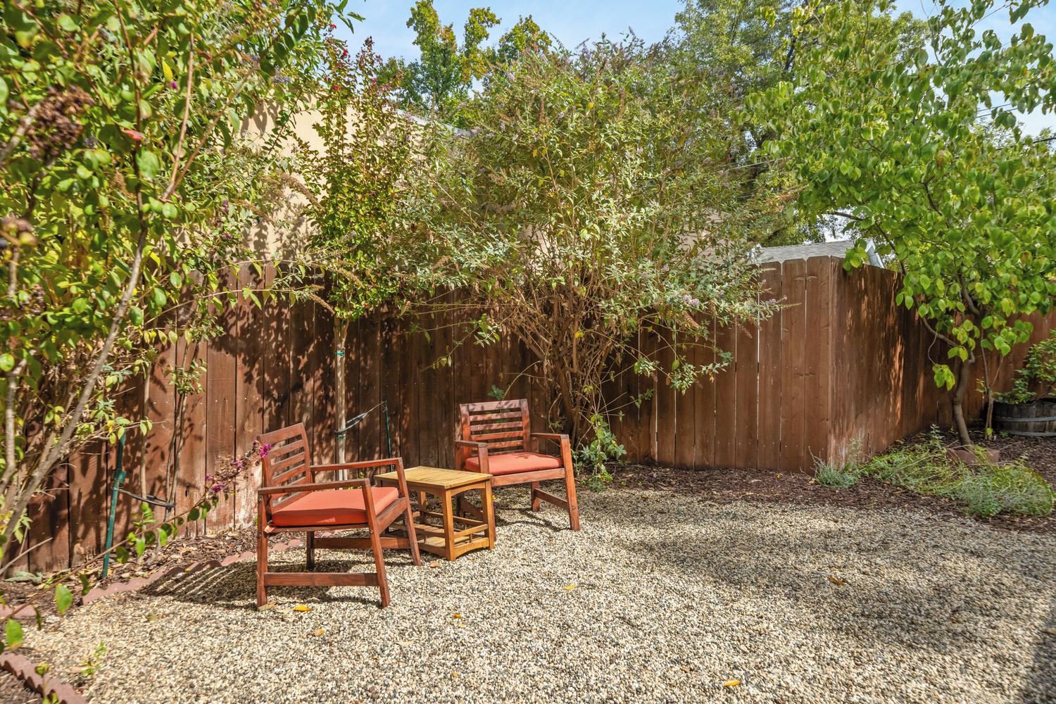 An outdoor seating area with two wooden chairs and a table, surrounded by a wooden fence and dense foliage.