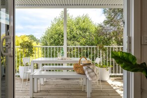 Une terrasse extérieure couverte avec une table et des chaises blanches, un panier, un chapeau et une plante en pot.