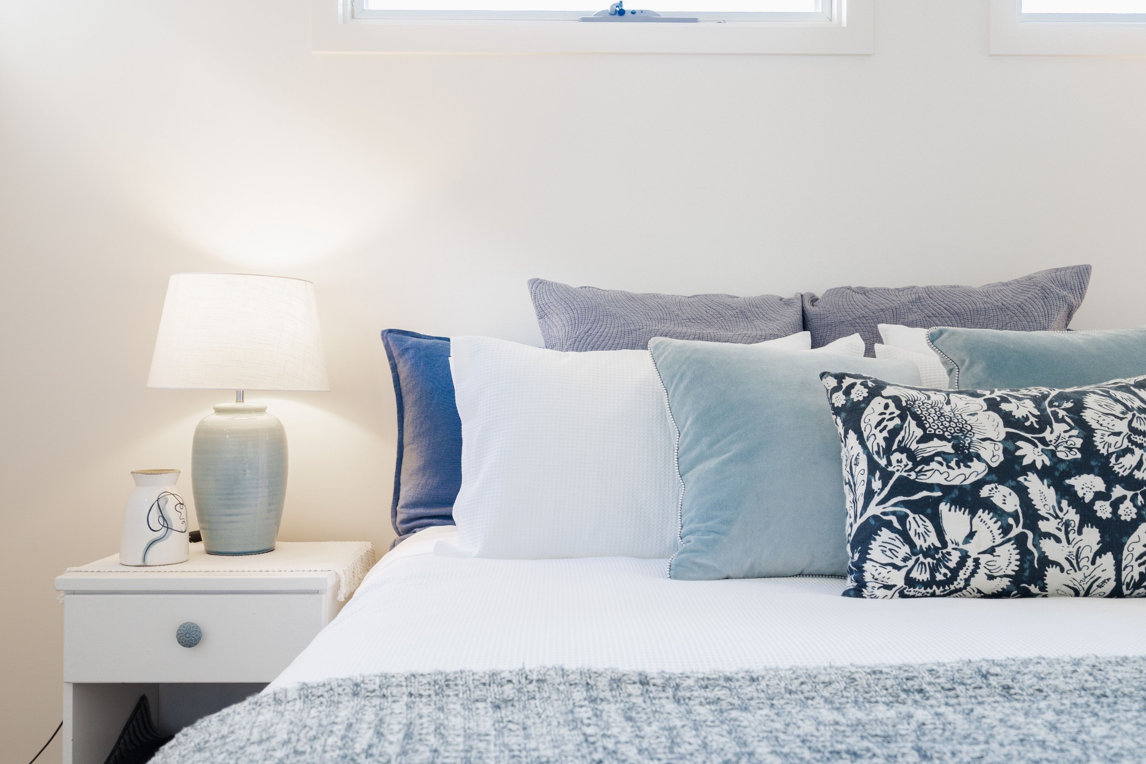Un lit bien fait, recouvert d’une couvre-lit blanche et bleue, agrémenté de plusieurs coussins, une lampe de chevet et un petit objet décoratif posé sur la table de chevet.