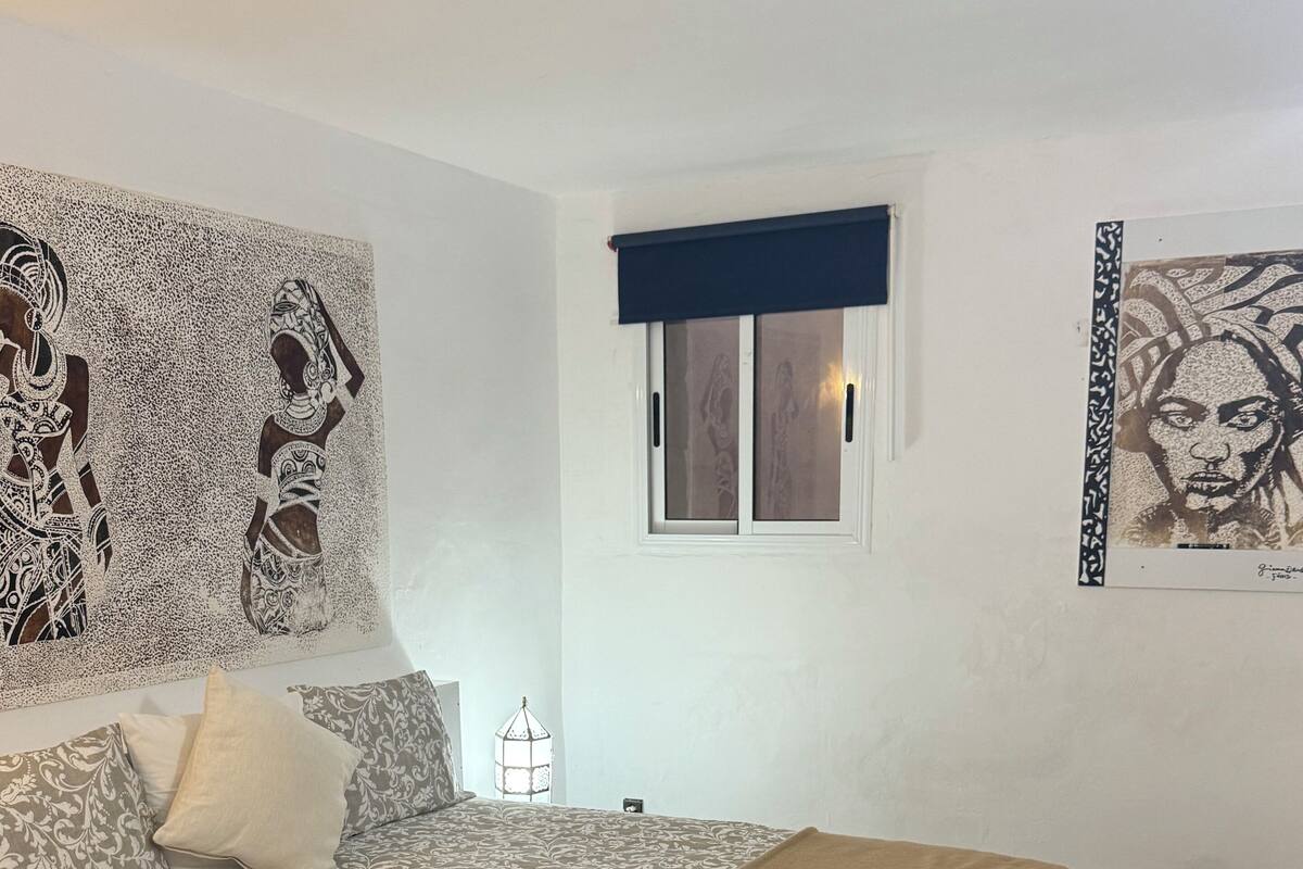 Ein Schlafzimmer mit einem Muster-Bettbezug, einer braunen Decke, einem Fenster mit blauem Vorhang und einer Wand mit einem großen abstrakten Kunstwerk.