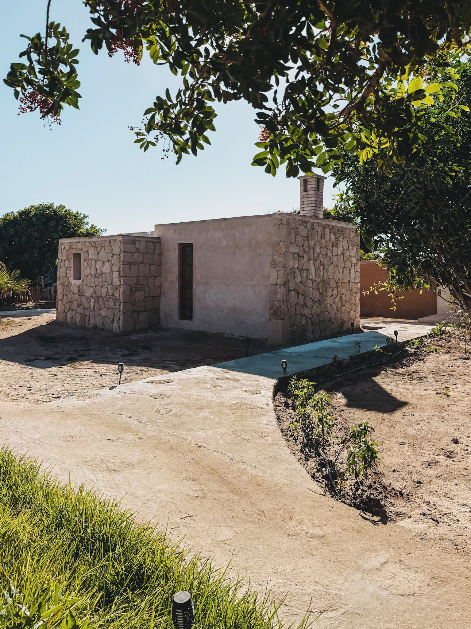 A rustic stone building with a door and a small window, surrounded by a sandy area and greenery.