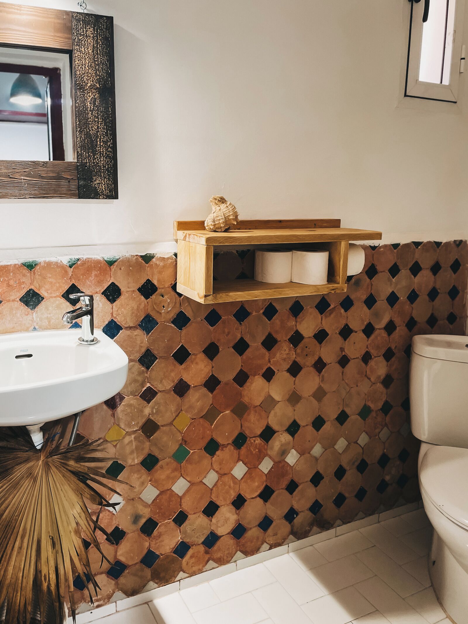 A bathroom with a mosaic tile wall, a wooden shelf with toiletries, a white sink, and a toilet.