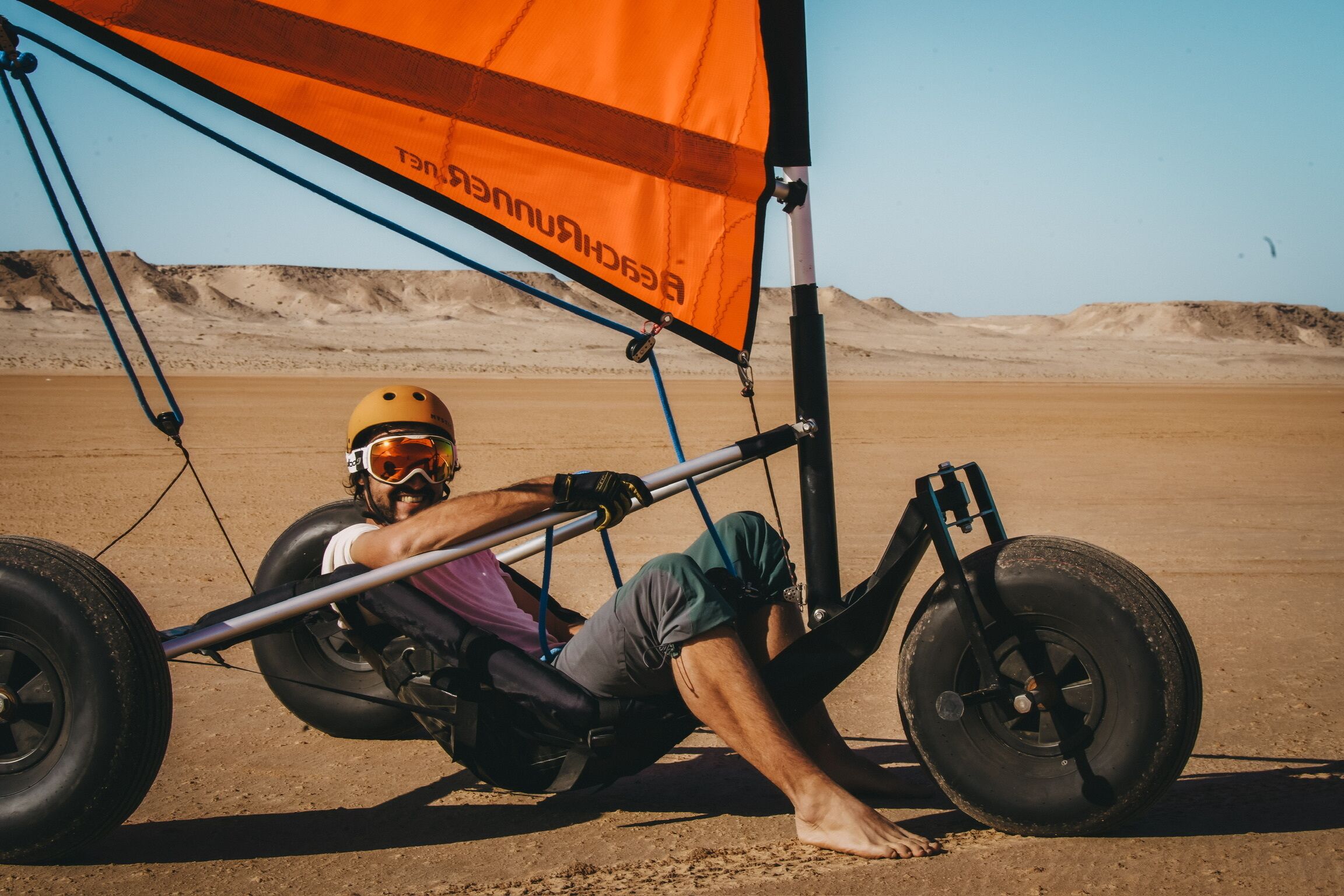A person in a desert setting, wearing a helmet and harness, attached to a glider with an orange canopy.