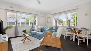 A modern living room with a blue sofa, a wooden rocking chair, a dining table with chairs, and large windows with curtains. - Jetty Rd Jewel, Top Location, Wi-Fi, Parking, Chic (Brighton)