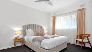 A neatly arranged hotel room with a large bed, a ceiling fan, a bedside table with a lamp, and a chair. - Jetty Rd Jewel, Top Location, Wi-Fi, Parking, Chic (Brighton)