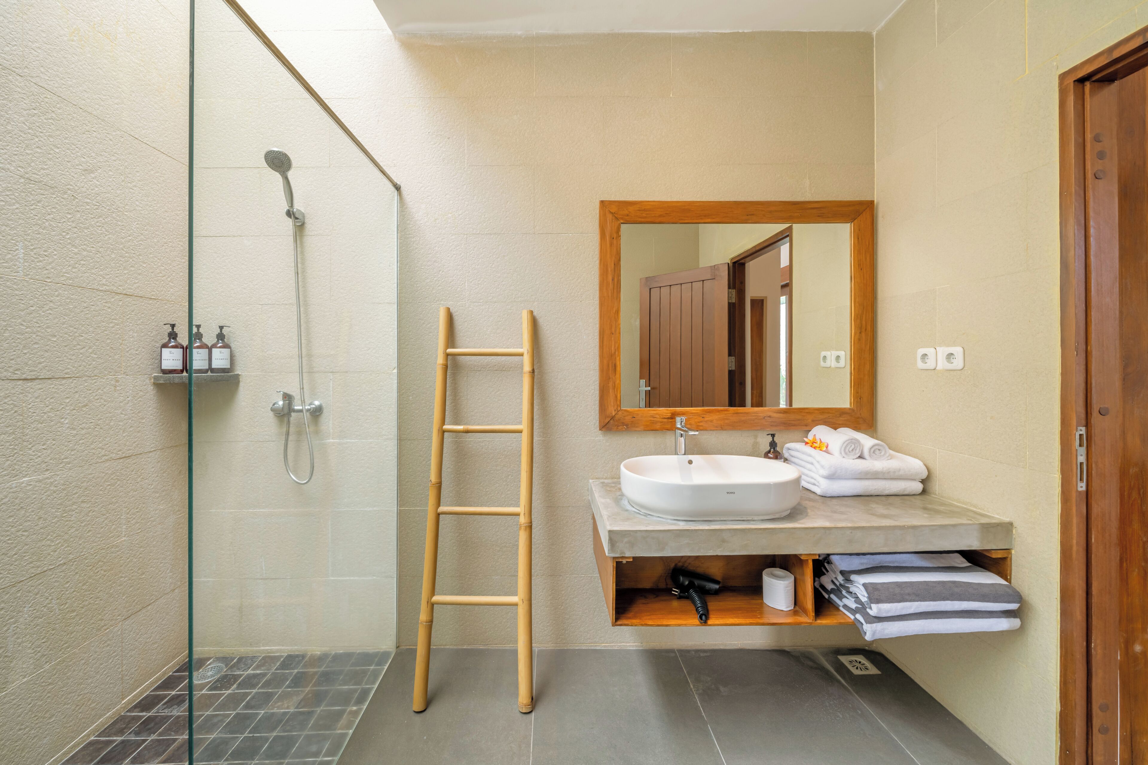 A modern bathroom with a glass shower enclosure, a wooden ladder, a large mirror, and a built-in sink with towels.