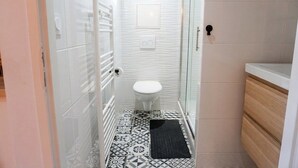 Casa de banho com sanita branca, pavimento em azulejos estampados, tapete preto e cabine de duche em vidro.
