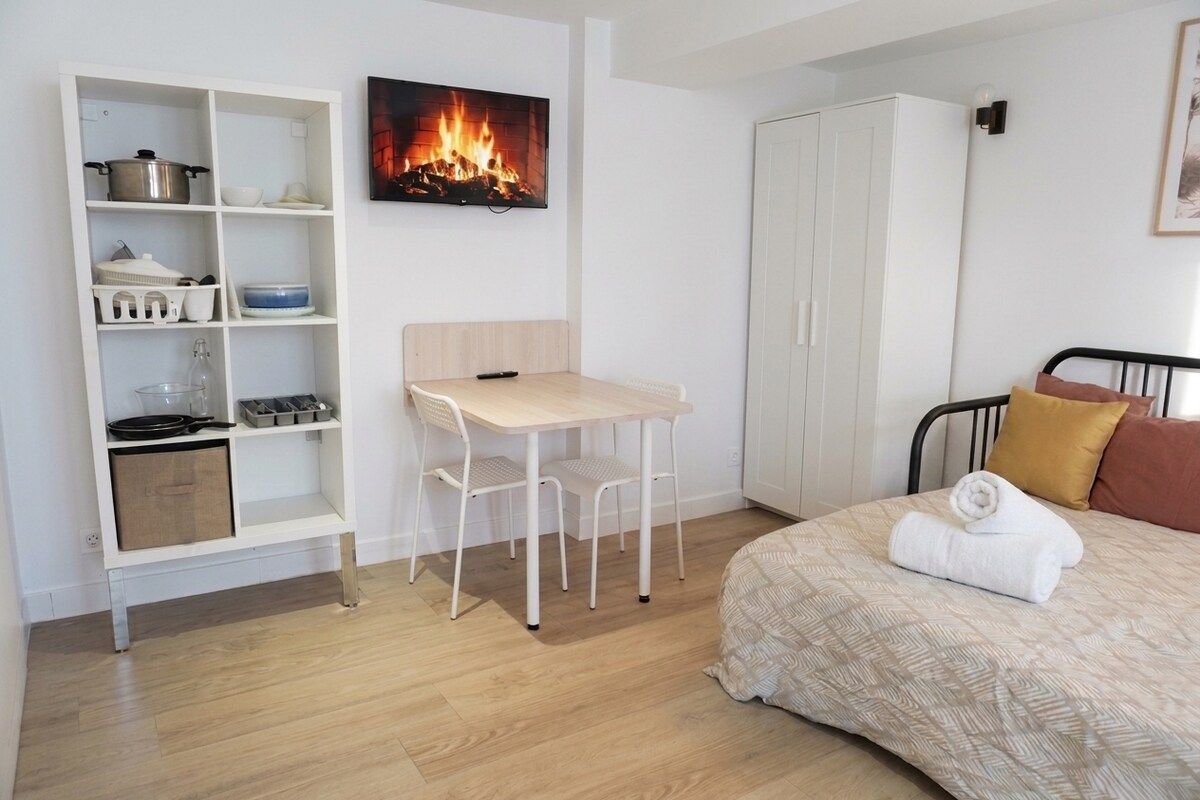 Een slaapkamer met een bed, nachtkastje, stoel, kledingkast en een wandplank met keukengerei.
