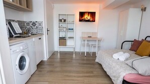 Quarto compacto com máquina de lavar, uma mesinha, cama e lareira na parede.