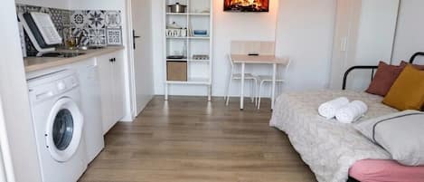 A compact room with a washing machine, a small table, a bed, and a wall-mounted fireplace.