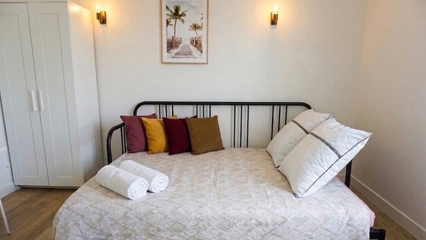Cama bem arrumada com colcha estampada e almofadas, guarda-roupa branco e um quadro pendurado na parede.