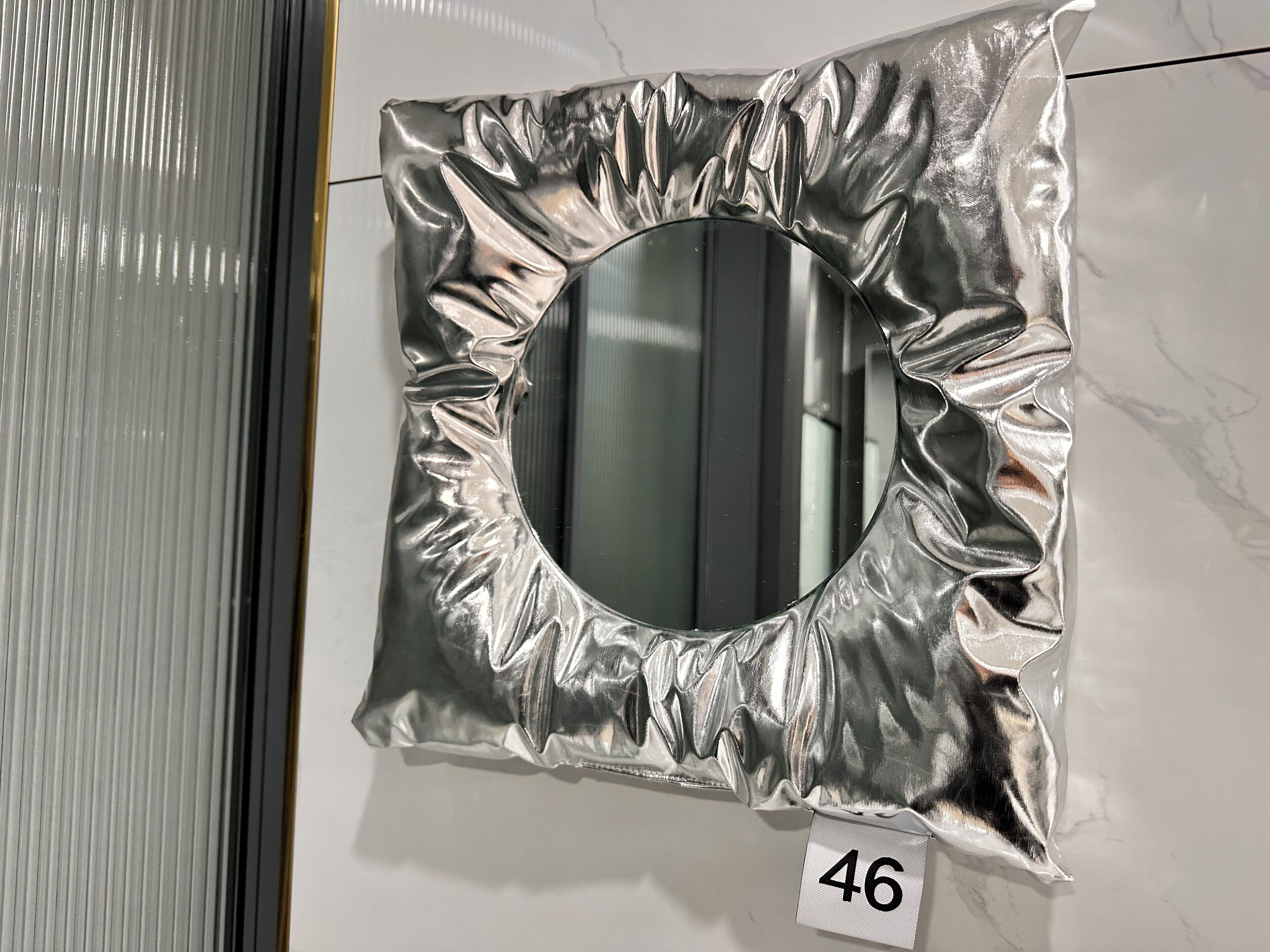 A metallic, crumpled mirror with a number tag '46' on a white wall.