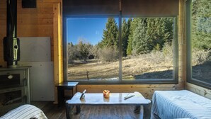 Una cabaña con un ventanal grande, una mesa de madera, dos camas, una estufa y un refrigerador.