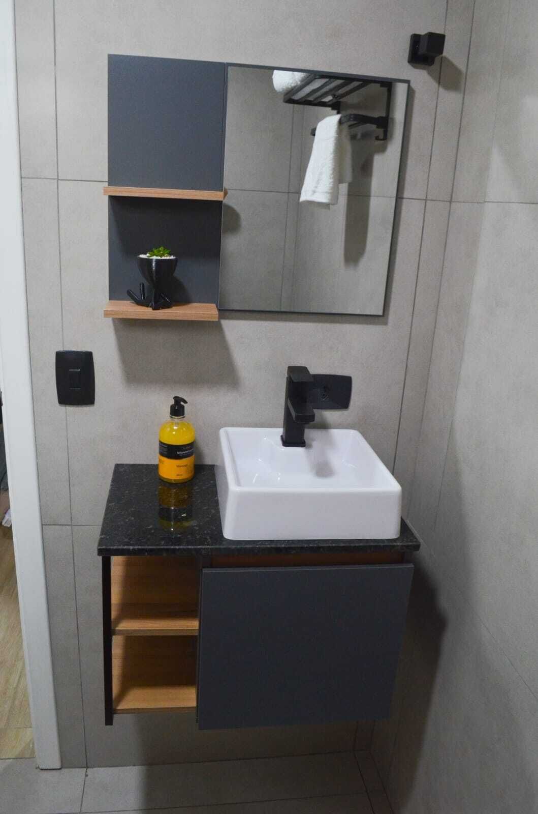 A modern bathroom with a white sink, black faucet, and a mirror above. There is a small shelf with a plant and a bottle of hand soap.