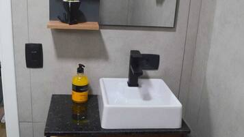 A modern bathroom with a white sink, black faucet, and a mirror above. There is a small shelf with a plant and a bottle of hand soap.