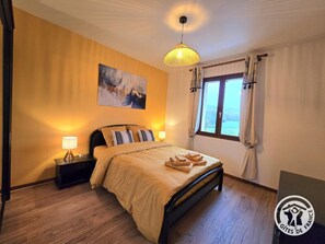 Ein Schlafzimmer mit Bett, Nachttischlampen, einem Gemälde, einem Fenster mit Vorhängen und einem Logo an der Wand.