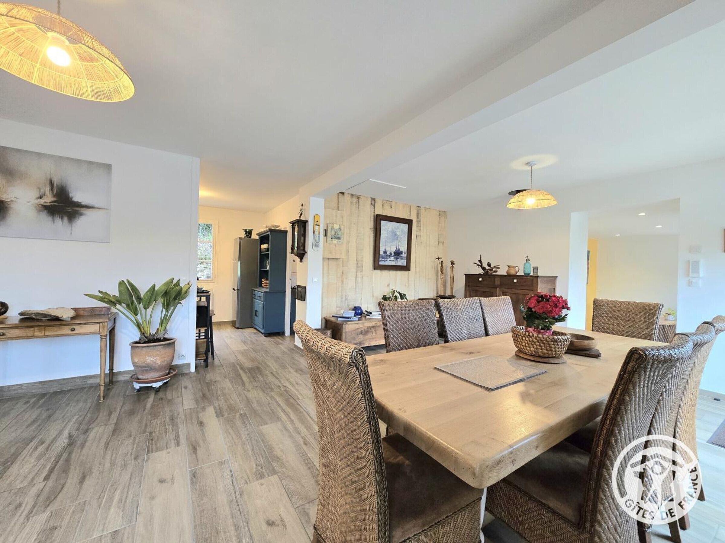 A spacious dining area with a wooden table and wicker chairs, a kitchen in the background, and a large window providing natural light.