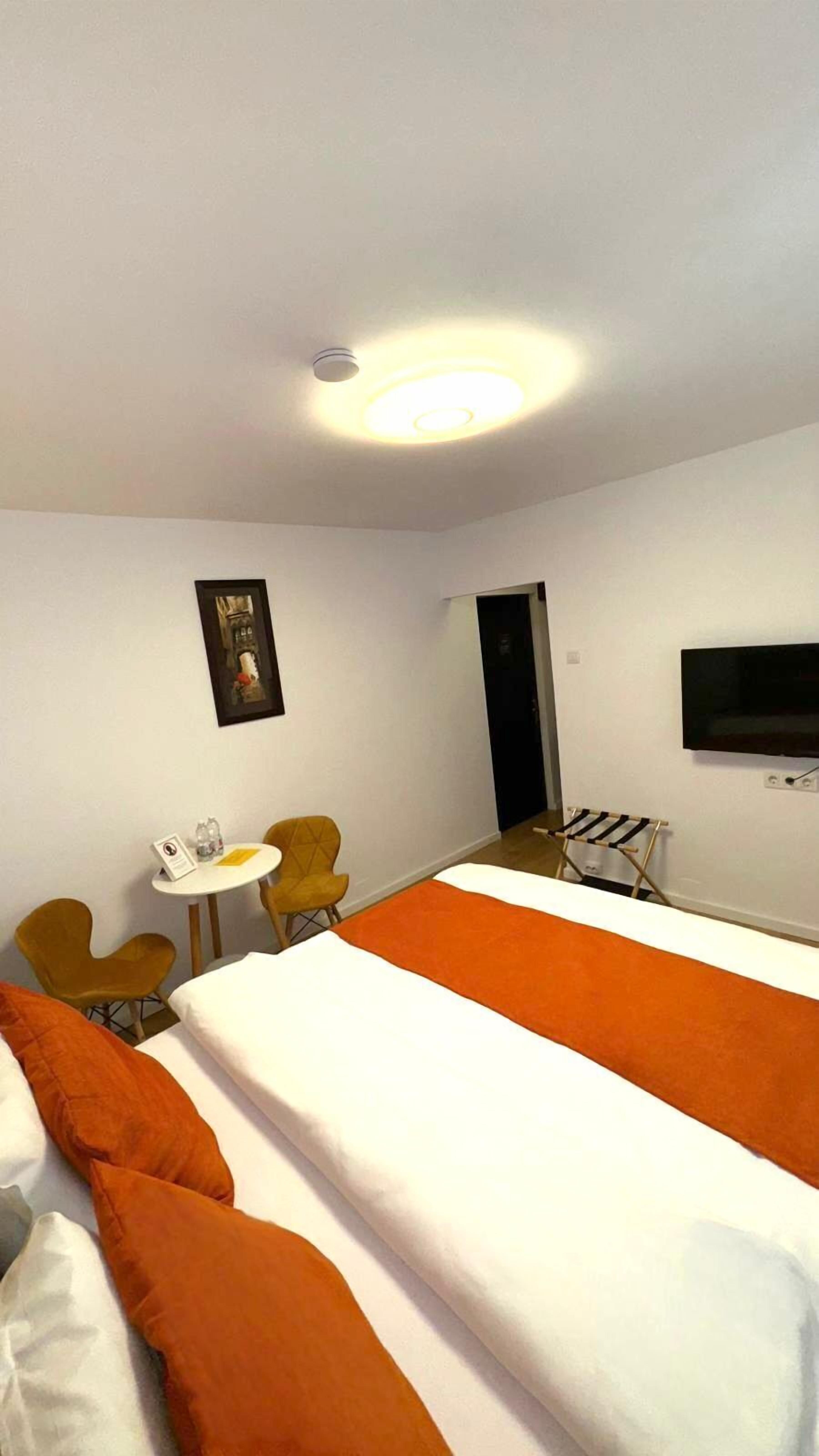 A hotel room with two beds, a small table with chairs, a TV mounted on the wall, and a ceiling light.